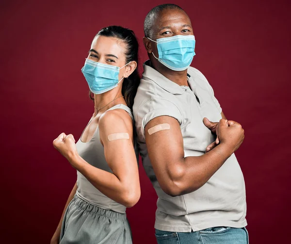 Covid vaccinated African american man and mixed race woman standing back to back. Two people wearing surgical face mask isolated against red background in studio with copyspace. Flexing plaster on ar.