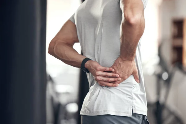 Closeup of one mixed race man holding his sore lower back while exercising in a gym. Guy suffering with painful spine injury from fractured joint and inflamed muscles during workout. Struggling with .
