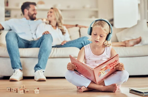 Little caucasian girl reading a story book while wearing headphones listening to music with her parents in the background. Child reading a fairytale and listening to music with her mom and dad at hom.
