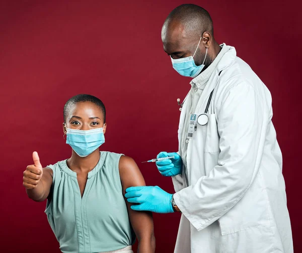 African american covid vaccinated woman showing thumbs up sign and symbol. Black doctor giving vaccine to patient wearing surgical face mask. Endorsing corona needle injection from physician in studi.