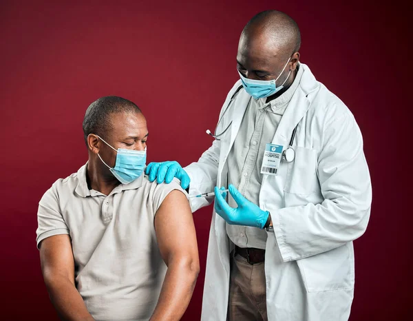 African american doctor giving covid vaccine to black man wearing surgical face mask. Healthy patient getting corona injection from physician with needle against red studio background with copyspace.
