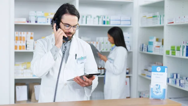 Pharmacist man answering the telephone and giving advice to customer on flu shot treatment options in pharmacy. Chemist assisting remote client by checking medicine stock on tablet.