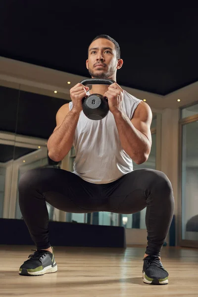 Its time to muscle up. Full length shot of a handsome young male athlete doing squats while holding a kettle bell in the gym