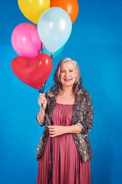 Never miss out on a chance to celebrate in life. Portrait of a cheerful senior woman posing holding balloons in studio against a blue background