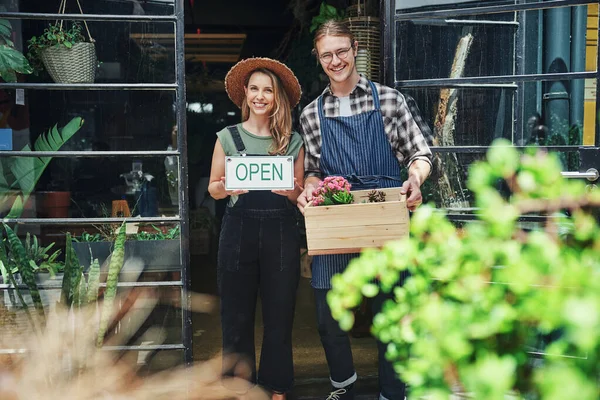 Come on in. Cropped portrait of two young business owners standing at the entrance of their floristry together and holding potting supplies