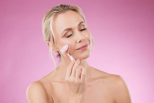 Beauty, facial roller and woman doing face massage with dermatology and cosmetics in studio. Mature aesthetic person on pink background for skincare, self care and rose quartz results for skin glow.