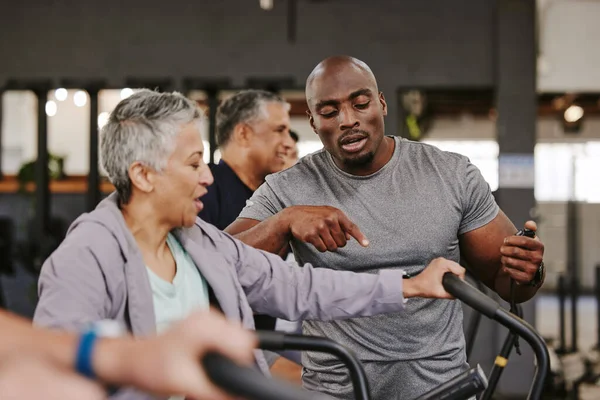 Elderly group, exercise bike and personal trainer for fitness, timer and retirement wellness by blurred background. Senior woman, bicycle training and diversity with black man, progress and coaching.