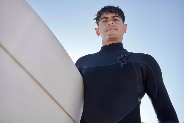 Surfing, beach and serious surfer with blue sky outdoor for water sports, training and exercise. Surfboard, morning workout and sea with summer sun and man ready for sport and freedom at the ocean.