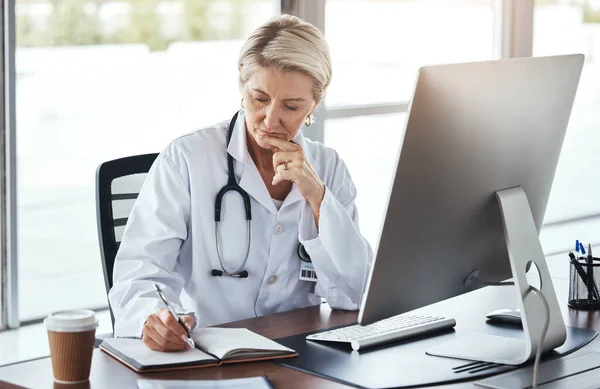 Senior woman, doctor and writing in book by office desk for healthcare research, notes or planning. Elderly female medical professional thinking with notebook by computer for health strategy or plan.