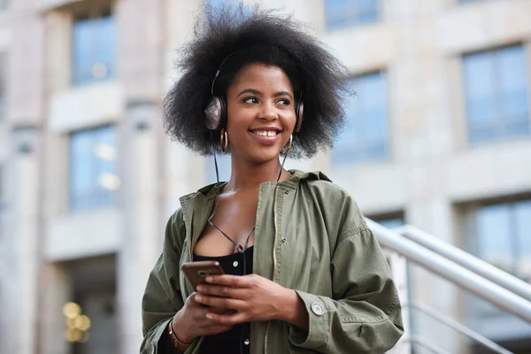 Music, phone and gen z black woman with headphones in city to relax and smile outside in street. Urban fashion, enjoying online streaming radio service and trendy girl listening to audio on earphones.