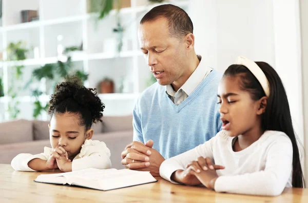 Family, worship and bible with father and kids praying at table for peace, religion and faith in their home. God, pray and children with parent for prayer, bible study or Jesus Christ praise together.