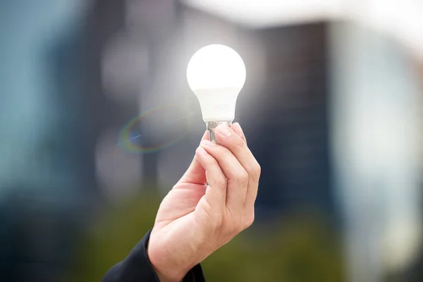 Hands, light bulb and idea in the city for solution, eco friendly or renewable energy on blurred background. Hand of person holding lamp for creativity, ideas or power saving element on mockup.