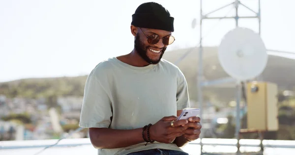 Phone, texting and happy black man on rooftop of city building using smartphone for social media, chatting and communication. Fashion, meme and young trendy guy typing message, text or post in town.