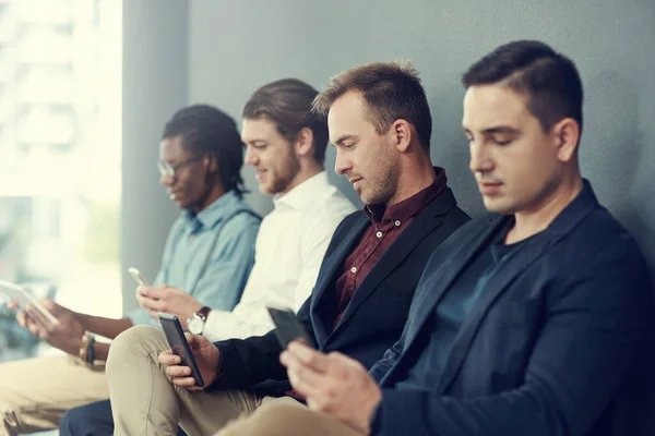 Their devices are keeping them busy while they wait. a group of businessmen using different wireless devices while waiting in line for an interview