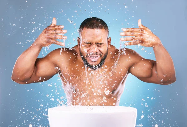 Water splash, hands or black man cleaning face for beauty, skincare or fresh hygiene on blue background. African male model with wellness washing, grooming or cleansing for facial treatment in studio.