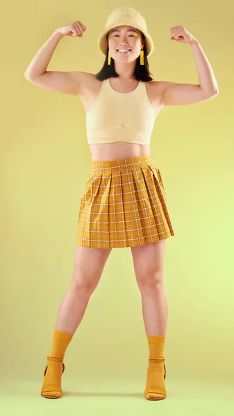 Smile, empowerment and asian woman flexing arms, full body portrait of gen z model with trendy fashion win in studio. Feminism, power and strength, happy and strong girl isolated on yellow background.