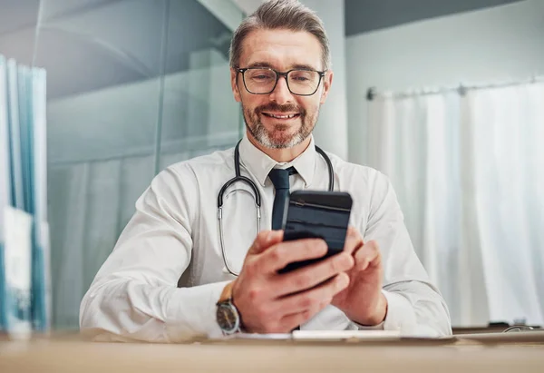 Healthcare, smile and doctor with smartphone at desk for wellness research, medical app and online consulting. Hospital, clinic and happy man with phone for internet, communication and telehealth.