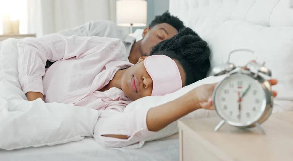 Black woman waking up in bed to switch off her alarm clock with a sleep mask in her house. Lazy, sleepy and African girl sleeping in her comfortable bedroom with an early morning get up time at home