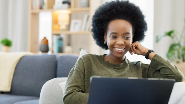 Laughing afro woman using laptop to watch funny, comedy movies or videos. Smiling, happy woman with stylish, funky and cool hair sitting alone on home living room sofa, relaxing and using technology.