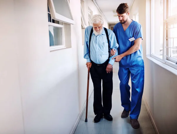 The road to recovery begins now. a young doctor helping his senior patient walk down a hallway in hospital