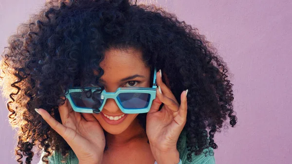 Black woman with sunglasses, smile, laugh and play with hair against pink backdrop in sunshine. Model girl with curly afro hair, happy with blue fashion glasses against pink wall or background.