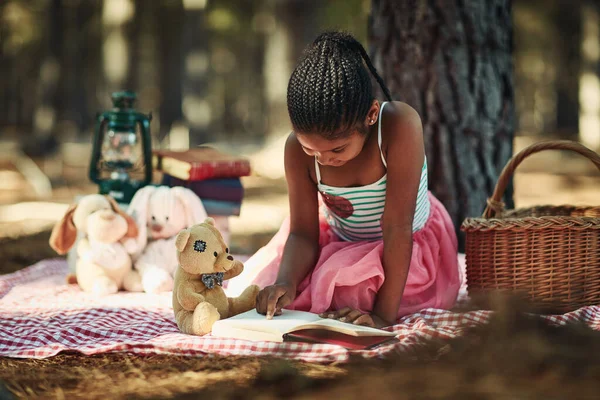 Learning and loving literature. a little girl reading a book with her toys in the woods