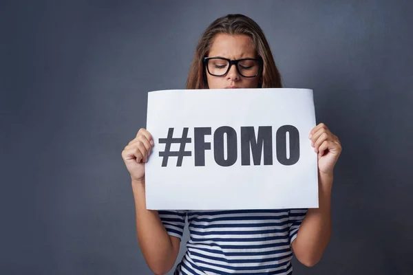 Am I missing out on something. Studio shot of an attractive young woman holding a sign with FOMO printed on it against a gray background