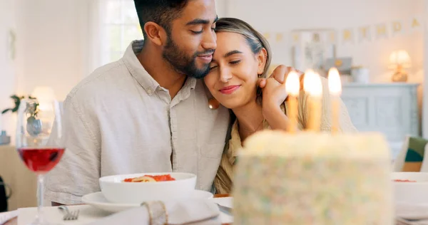 Interracial couple, gift and celebrate birthday being happy, kiss and smile in home at table with cake. Love, man and woman being content, romantic and present being cheerful celebration together