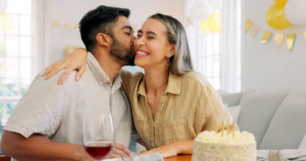 Interracial couple, gift and celebrate birthday being happy, kiss and smile in home at table with cake. Love, man and woman being content, romantic and present being cheerful celebration together