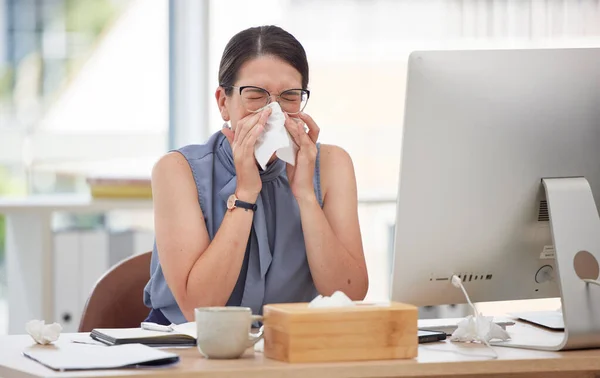 Sick, computer and blowing nose with woman in office for virus, illness and allergy symptoms. Sneezing, disease and tissue with employee suffering with infection at desk for flu, fever and cold.