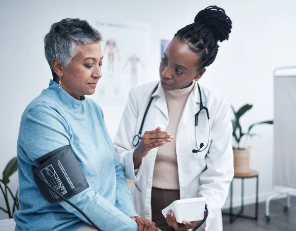 Black woman, doctor and senior patient with blood pressure reading for wellness, advice and conversation. Medic, elderly client and medical tools for health, cardiology and results in hospital office.