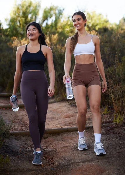 Fitness women or friends walking in nature with water bottle for workout, running and cardio training together. Diversity athlete, sports or runner people in woods with diet, body and wellness goals.