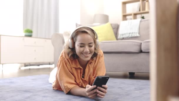 ヘッドフォン 音楽を持つアジアの女性は 心の健康のためのラウンジでカーペットの上でリラックスします 携帯電話 スマートフォン モバイルアプリの音声技術を聴くリビングルームの床の上の人 — ストック動画