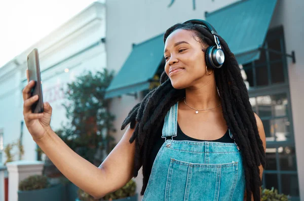 Phone, selfie and happy black woman in the city street with headphones listening to music, podcast or radio. Happiness, technology and african girl on a walk in the urban road on a smartphone