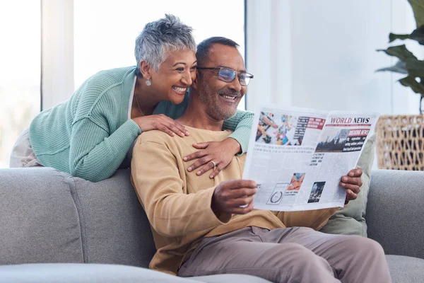 Love, senior couple and reading newspaper, story or news article in living room home. Retirement, relax time and happy elderly man and woman embrace in house, hugging or cuddle together and bonding