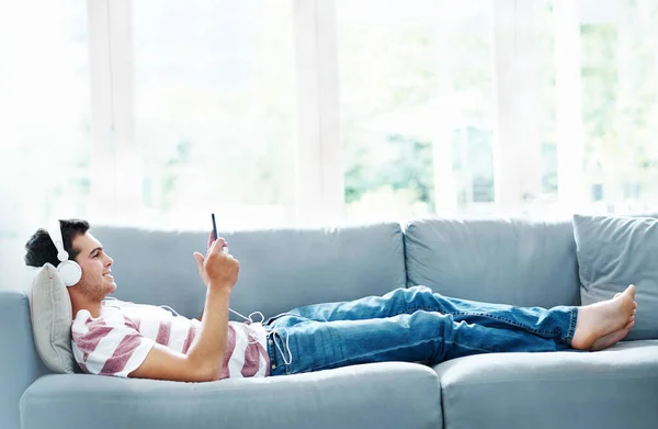 新しいタブレットを楽しんでる 家のソファに横になって音楽を聴く魅力的な男 — ストック写真