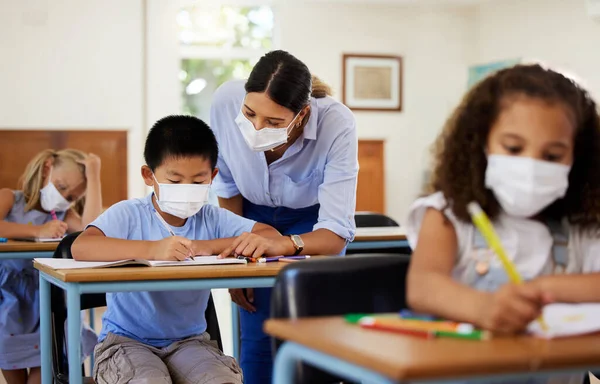 Covid, education and learning with a teacher wearing a mask and helping a male student in class during school. Young boy studying in a classroom with help from an educator while sitting at his desk.