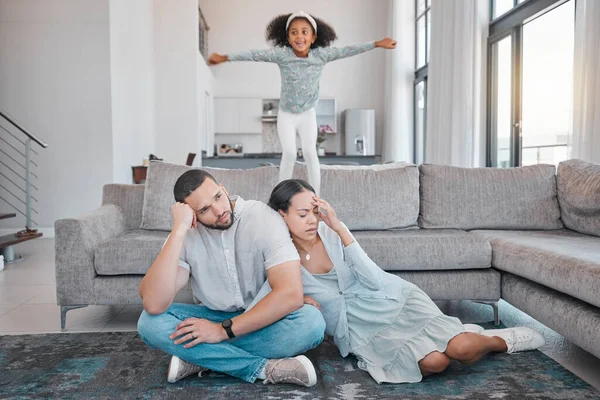 Tired, parents and child jump on sofa with exhausted mother and father sitting on floor in living room. Family, energy and young girl jumping on couch with frustrated, annoyed and upset mom and dad.