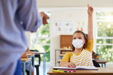 Smart student with covid face mask asking teacher question about corona virus pandemic in a classroom or elementary school. Little girl child raising hand to answer healthcare related topic in class.