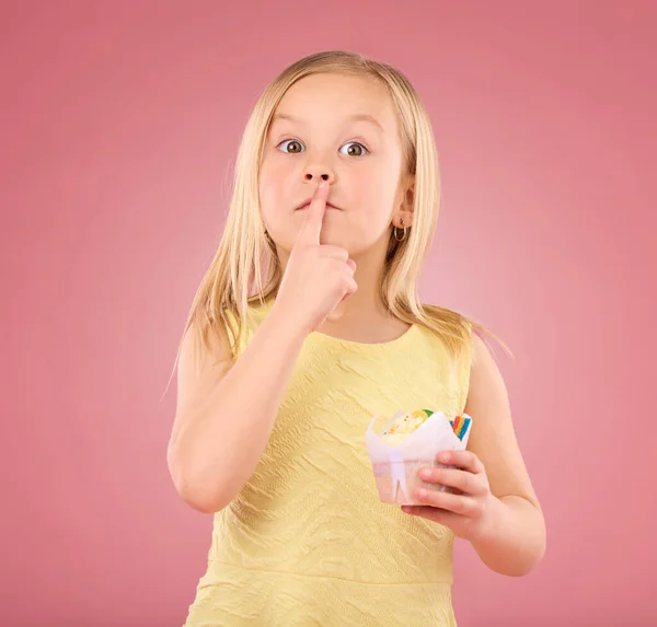 Girl child, finger on lips and cupcake portrait in studio on a pink background for hush and silence. Face of female kid model with cake, secret and sweet snack in hand isolated on color and space.