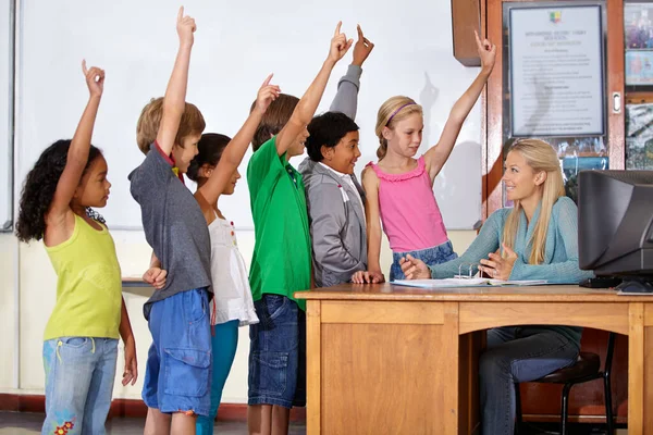 教书是她的梦想 一位老师坐在课桌前与她的学生们交谈 他们个个都把手举起来 — 图库照片