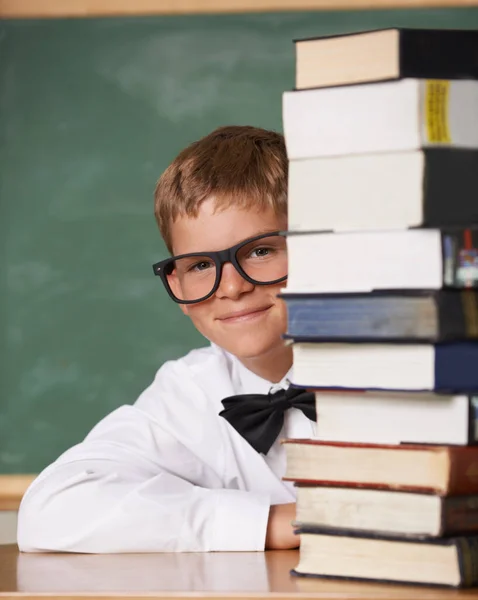 He has a thirst for knowledge. A young boy wearing glasses and a bow-tie smiling at the camera from behind a stack of books