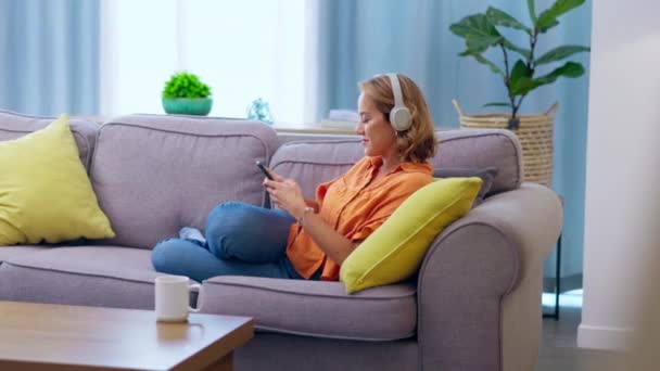 沙发上的女人 在家里客厅里玩乐 放松快乐的女性 拥有流动的智能手机 喜欢播客 收音机或音频歌曲 — 图库视频影像