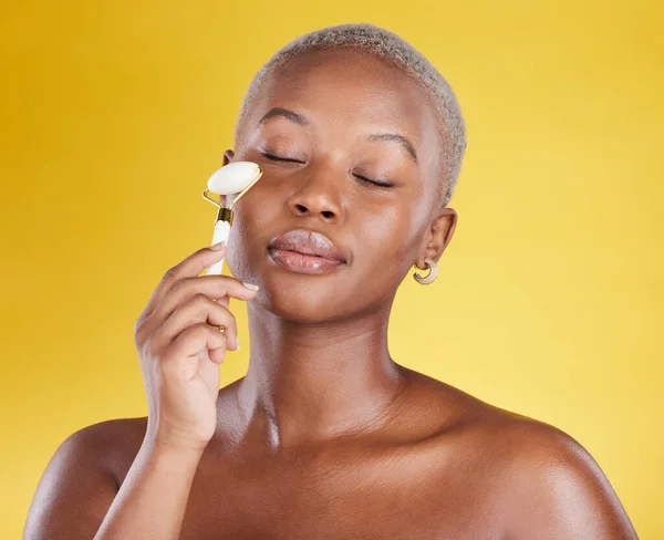 Face, beauty skincare and black woman with roller in studio isolated on a yellow background. Dermatology health, massage and African female model with eyes closed and stone for facial skin treatment