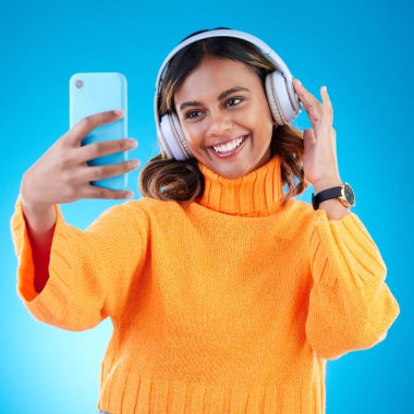 Müzik kulaklıkları, gülücük ve stüdyoda mavi arka planda izole edilmiş bir kadının selfie 'si. Profil fotoğrafı, radyo ve mutlu Hintli kadın yayını, dinleme ve keyif alma sesi, ses podcastı veya şarkı