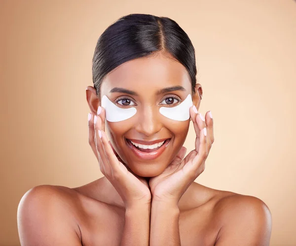 Beauty, eye patch and portrait of a woman in studio for skincare, dermatology and cosmetics. Happy Indian female model with smile and collagen mask for self care moisture facial on brown background.