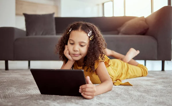 Full length adorable little mixed race child using a digital tablet while thinking at home. One small cute hispanic girl lying alone on living room floor and playing a game on technology. Bored kid.
