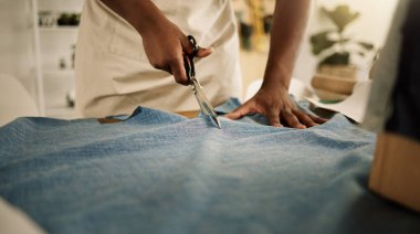 Yaratıcı tasarımcı kumaşını kesiyor. Makas kullanarak kot kumaşı kesen bir terzinin yakın çekimi. Moda tasarımcısı stüdyosunda çalışıyor. Girişimci tekstil ürünlerini bir masada kesiyor..