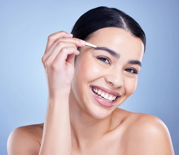 Studio portrait of a smiling mixed race young woman with glowing skin posing against blue copyspace background while tweezing her eyebrows. Hispanic model using a tweezer for hair removal.