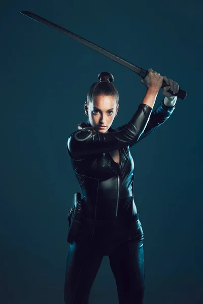 Woman, warrior and vigilante in cosplay with sword ready for battle, war or game against a dark studio background. Female in black costume or hero with guns and blade for halloween or super power.